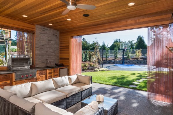 Covered patio behind new luxury home with fireplace, tv, and couch, with view of lush green grass and landscaping